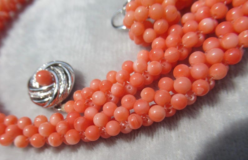 Op de afbeelding: Een meerstrengs ketting bestaande uit kleine, ronde, zalmkleurige kralen. De ketting heeft een ronde, zilverkleurige metalen sluiting met een gestructureerd, wervelend ontwerp en een bijpassende zalmkleurige kraal in het midden.