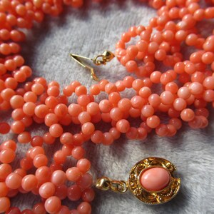 Puede incluir: Un collar de varias hebras con peque&ntilde;as cuentas redondas de color coral. El collar tiene un cierre dorado y un colgante decorativo con una piedra de color coral. Las cuentas est&aacute;n dispuestas en m&uacute;ltiples hebras.