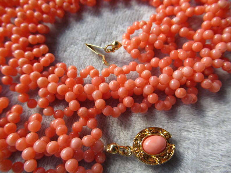 Puede incluir: Un collar de varias hebras con peque&ntilde;as cuentas redondas de color coral. El collar tiene un cierre dorado y un colgante decorativo con una piedra de color coral. Las cuentas est&aacute;n dispuestas en m&uacute;ltiples hebras.