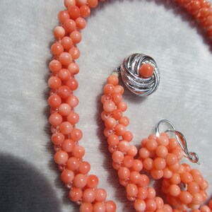 Op de afbeelding: Een ketting bestaande uit meerdere strengen kleine, ronde, zalmkleurige kralen die met elkaar zijn verweven. De ketting heeft een ronde, zilverkleurige sluiting met een gestructureerd wervelend ontwerp en een bijpassende kraal in het midden.