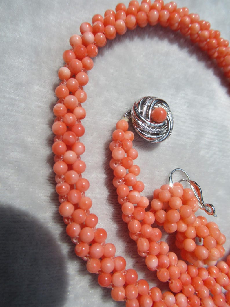 Op de afbeelding: Een ketting bestaande uit meerdere strengen kleine, ronde, zalmkleurige kralen die met elkaar zijn verweven. De ketting heeft een ronde, zilverkleurige sluiting met een gestructureerd wervelend ontwerp en een bijpassende kraal in het midden.