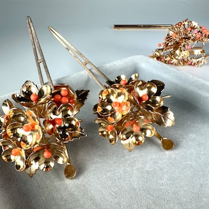 Op de afbeelding: Twee gouden haarstokken met bloemmotieven en koraalkleurige kralen. De bloemen zijn van metaal en hebben een delicaat, ingewikkeld ontwerp.