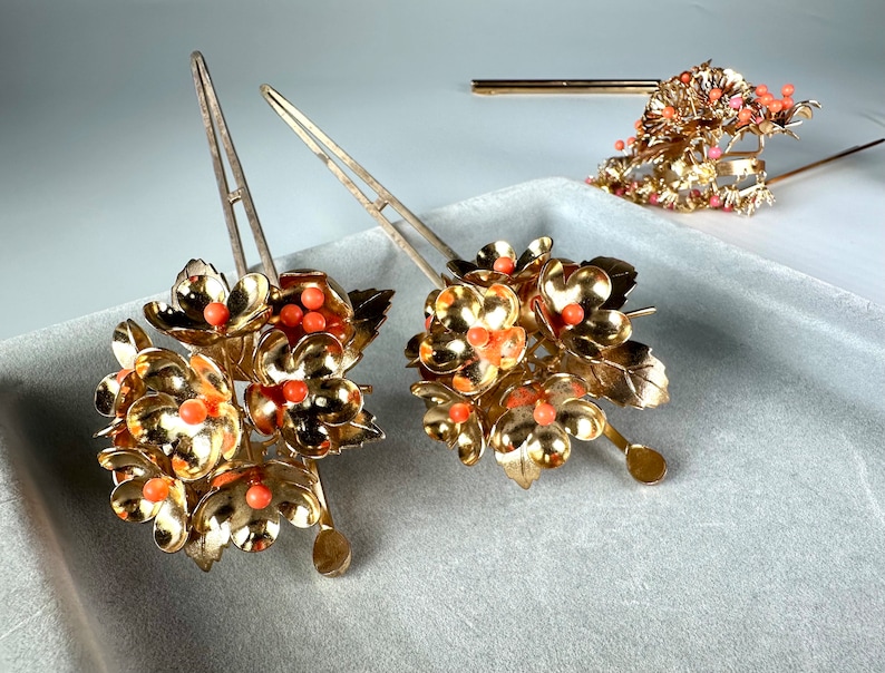 Op de afbeelding: Twee gouden haarstokken met bloemmotieven en koraalkleurige kralen. De bloemen zijn van metaal en hebben een delicaat, ingewikkeld ontwerp.