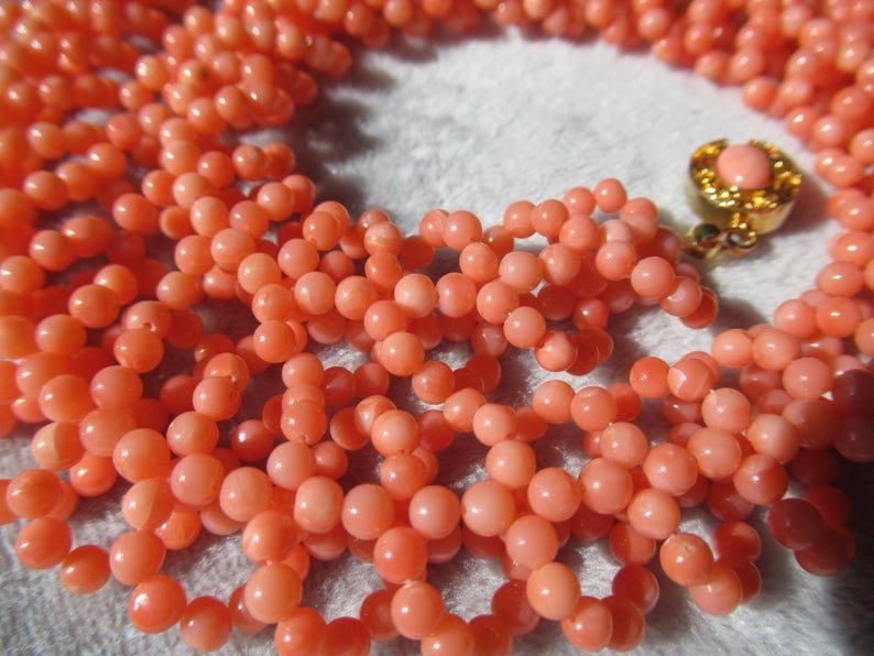 Puede incluir: Primer plano de un collar de cuentas de coral. El collar presenta m&uacute;ltiples hilos de peque&ntilde;as cuentas redondas de color melocot&oacute;n. Se ve un cierre dorado con una piedra de color melocot&oacute;n. Las cuentas son de tama&ntilde;o uniforme y est&aacute;n bien ensartadas.