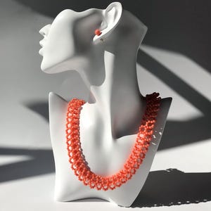 Puede incluir: Un collar de cuentas de color coral y pendientes a juego exhibidos en una cabeza de maniqu&iacute; blanca. El collar es un dise&ntilde;o de m&uacute;ltiples hilos, y los pendientes son peque&ntilde;as cuentas redondas. La joyer&iacute;a se encuentra sobre un fondo blanco con sombras.