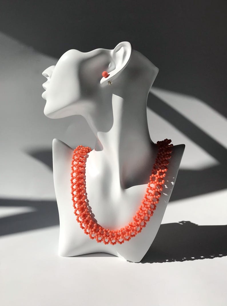 Puede incluir: Un collar de cuentas de color coral y pendientes a juego exhibidos en una cabeza de maniqu&iacute; blanca. El collar es un dise&ntilde;o de m&uacute;ltiples hilos, y los pendientes son peque&ntilde;as cuentas redondas. La joyer&iacute;a se encuentra sobre un fondo blanco con sombras.