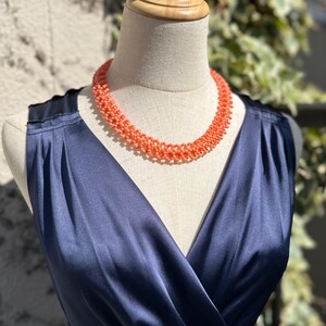 Puede incluir: Un collar de cuentas de color coral se exhibe en un maniqu&iacute;. El collar est&aacute; drapeado sobre un vestido de sat&eacute;n azul marino con cuello en V. El vestido es sin mangas y tiene un corpi&ntilde;o ajustado.
