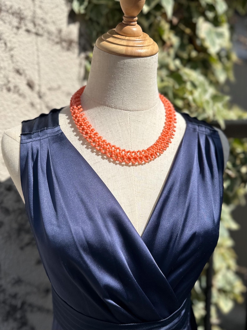 Puede incluir: Un collar de cuentas de color coral se exhibe en un maniqu&iacute;. El collar est&aacute; drapeado sobre un vestido de sat&eacute;n azul marino con cuello en V. El vestido es sin mangas y tiene un corpi&ntilde;o ajustado.