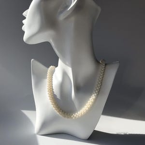May include: A white mannequin wearing a white pearl necklace. The necklace is made of small, round pearls.