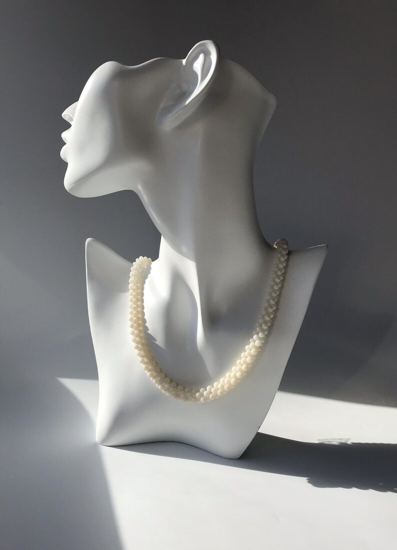 May include: A white mannequin wearing a white pearl necklace. The necklace is made of small, round pearls.
