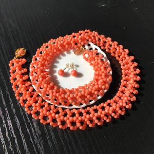 Puede incluir: Un collar de cuentas de color coral y pendientes a juego se exhiben en un plato blanco. El collar est&aacute; dispuesto en forma circular y los pendientes tienen una peque&ntilde;a cuenta redonda de coral. Las joyas se colocan sobre un fondo oscuro.