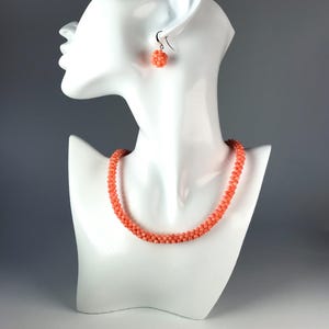 Op de afbeelding: Een koraalkleurige kralenketting en een bijpassende enkele kralenoorbel. De ketting bestaat uit een doorlopende streng van kleine, ronde, getextureerde kralen in een levendige zalmtint. De oorbel bevat een bolvormige tros van kralen.