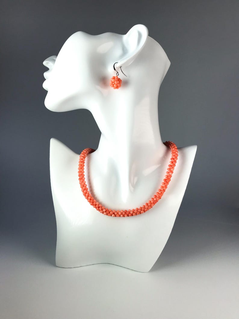 Op de afbeelding: Een koraalkleurige kralenketting en een bijpassende enkele kralenoorbel. De ketting bestaat uit een doorlopende streng van kleine, ronde, getextureerde kralen in een levendige zalmtint. De oorbel bevat een bolvormige tros van kralen.