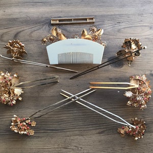 Op de afbeelding: Een set van zeven gouden haar kammen en pinnen met bloem- en bladmotieven. De kammen hebben rode en witte kralen. De pinnen zijn lang en dun met een eenvoudig ontwerp.