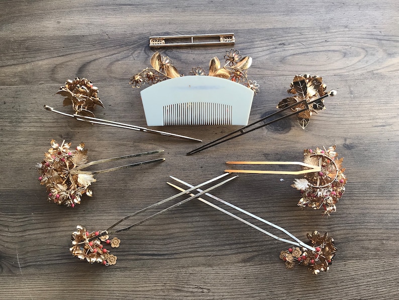 Op de afbeelding: Een set van zeven gouden haar kammen en pinnen met bloem- en bladmotieven. De kammen hebben rode en witte kralen. De pinnen zijn lang en dun met een eenvoudig ontwerp.