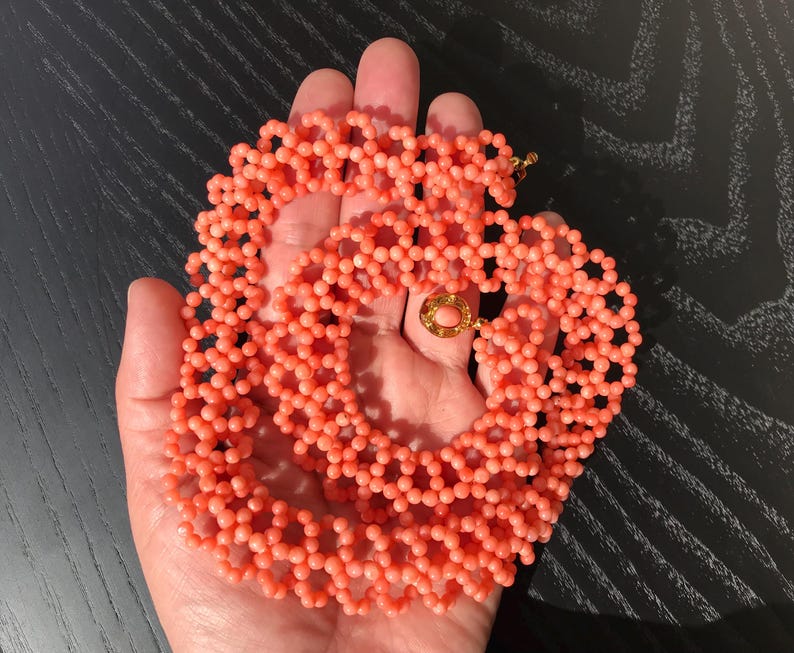 Puede incluir: Un collar de cuentas de color coral con un cierre dorado. El collar est&aacute; hecho de peque&ntilde;as cuentas redondas dispuestas en un patr&oacute;n delicado. El cierre es redondo con una piedra de color coral en el centro.