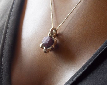 May include: A gold chain necklace with a pendant featuring a dark red gemstone. The pendant is made of gold and has a geometric shape. The necklace is worn by a person with a dark shirt.