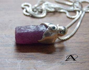Puede incluir: Un collar con un colgante de plata, adornado con una gran piedra preciosa púrpura en bruto engastada en una caja de plata. Dos pequeñas piedras preciosas verdes están engastadas en la caja de plata. El collar está en una cadena de plata.
