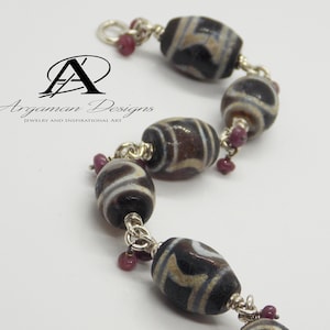 Könnte beinhalten: Ein silbernes Armband mit dunkelbraunen und weißen Perlen mit Mustern und kleinen rosafarbenen Edelsteinen. Das Armband ist mit einer silbernen Kette und einem Verschluss versehen.