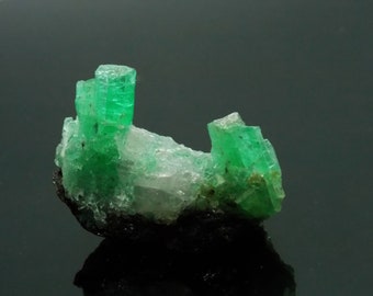 Può includere: Un gruppo di cristalli verde smeraldo su una roccia grigio scuro. I cristalli sono nitidi e ben definiti, con una lucentezza vitrea.
