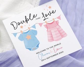 May include: A white card with the text "Double the Love" and a string of flags with a blue baby onesie and a pink baby dress hanging from it. The text "Welcome to the world Oliver & Esme 25th September 2024" is also on the card.