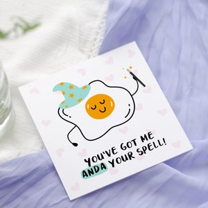 May include: A white greeting card with a cartoon fried egg wearing a wizard hat and holding a wand. The card has the text "YOU'VE GOT ME ANDA YOUR SPELL!" and small pink hearts.