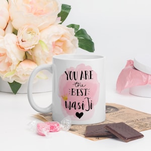 Puede incluir: Taza de cerámica blanca con el texto "YOU ARE the BEST Masi Ji" en escritura negra y un fondo rosa de acuarela. Un corazón negro está debajo del texto. La taza está junto a un ramo de flores rosas y chocolate.