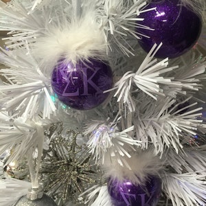 May include: Close-up of a white Christmas tree decorated with purple glitter ornaments. Each ornament has a white feather accent and purple Greek letters. Additional silver and glitter ornaments are also visible.