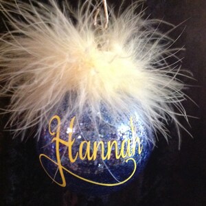 May include: A blue glass Christmas ornament with a white feather trim and the name "Hannah" written in yellow glitter.