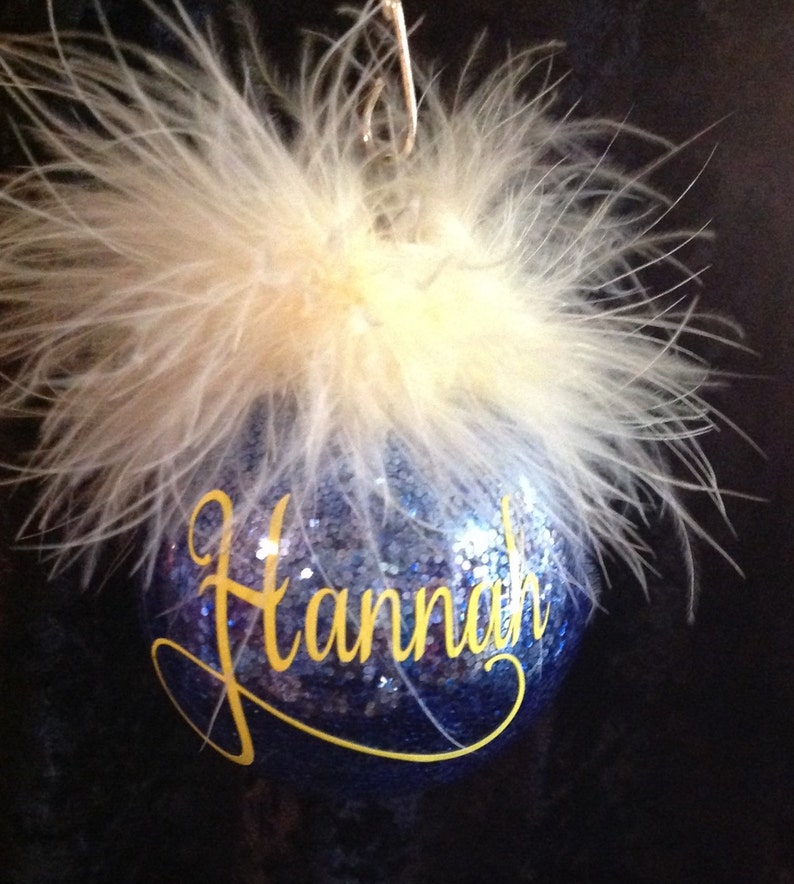 May include: A blue glass Christmas ornament with a white feather trim and the name "Hannah" written in yellow glitter.