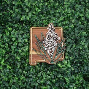 May include: Wooden cutout of the state of New Mexico with a white yucca flower design in the center. The state outline is brown and the flower is white with teal green leaves.