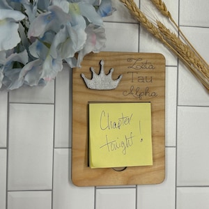 May include: A wooden sign with the text "Zeta Tau Alpha" and a silver crown. A yellow sticky note is attached to the sign with the text "Chapter tonight!"