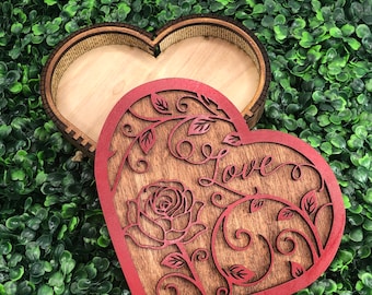 May include: A wooden heart-shaped box with a laser-cut lid. The lid is red and features a rose and the word "Love" in cursive. The box is open, revealing a natural wood interior. The box is on a green background.