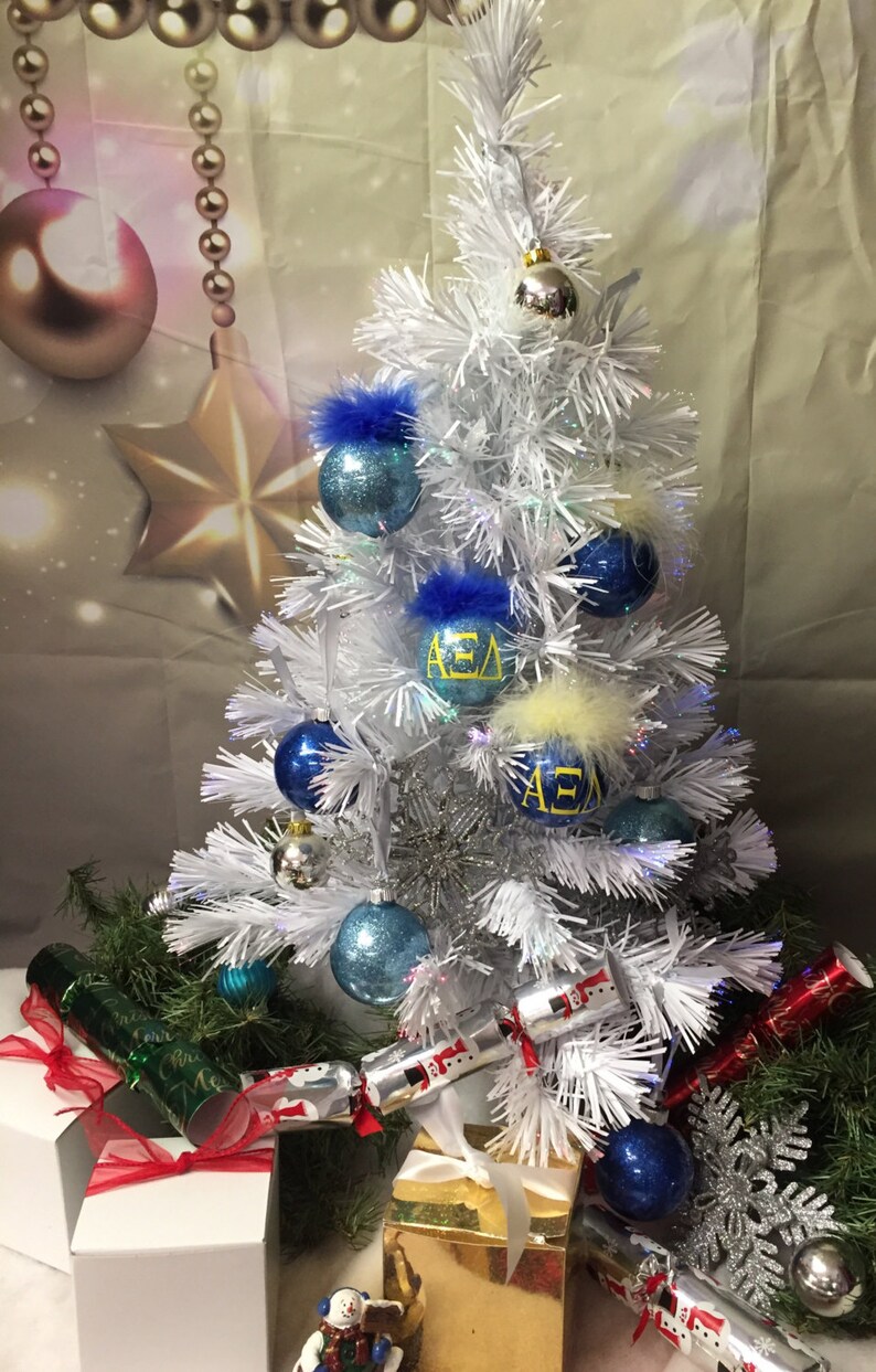 May include: A white artificial Christmas tree decorated with blue and silver ornaments, some with the letters "AEA" in yellow. The tree is surrounded by wrapped gifts and Christmas crackers.