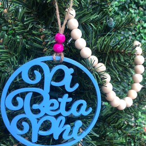 May include: A blue and white Christmas ornament with the text "Alpha Beta Phi" in a decorative script font. The ornament is hanging from a string of wooden beads with pink accents.
