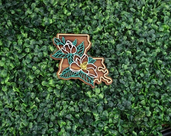 May include: A wooden cutout of the state of Louisiana with a floral design. The outline of the state is brown, the flowers are white, and the leaves are green.