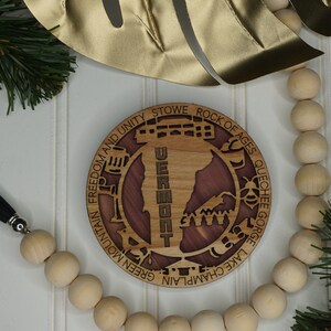 May include: A round wooden ornament featuring a map of Vermont with the state name and various landmarks and locations inscribed around the edge. The ornament is made of light and dark wood and has a natural wood finish.