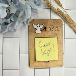 May include: A wooden sign with the text "Pi Beta Phi" and a small white angel figurine. A yellow sticky note is attached to the sign with the text "Chapter tonight!"