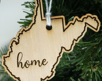 May include: Wooden ornament shaped like the state of West Virginia with the word "home" inscribed on it. The ornament is hanging from a white string on a green Christmas tree.