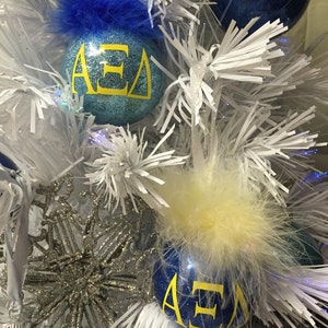May include: Two blue Christmas ornaments with white glitter and yellow lettering that reads "&Alpha;&Epsilon;&Delta;". One ornament has a blue feather trim and the other has a yellow feather trim.