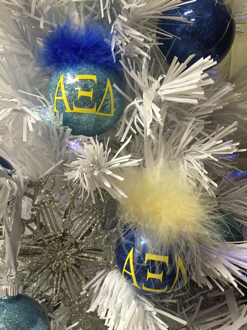 May include: Two blue Christmas ornaments with white glitter and yellow lettering that reads "&Alpha;&Epsilon;&Delta;". One ornament has a blue feather trim and the other has a yellow feather trim.