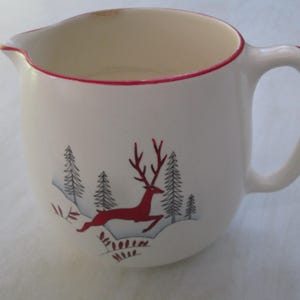 May include: Cream ceramic creamer with a red rim and handle. The creamer features a red deer leaping through a winter scene with grey and black trees. A charming addition to any kitchen or dining set.