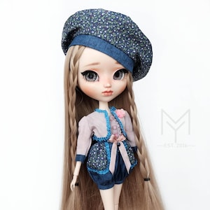 May include: A doll with long blonde hair styled in braids, wearing a blue and white floral patterned beret, a light pink jacket with a blue and white floral trim, and blue denim shorts with a pink ribbon.