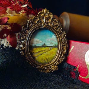 Op de afbeelding: Een kleine, sierlijke, goudkleurige lijst met een miniatuurlandschap van een veld en lucht. De lijst rust op een rood boek met gouden letters, naast gedroogde rode en gele bloemen. De achtergrond is donker.