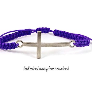 Puede incluir: Pulsera de cordón trenzado morado con un dije de cruz plateada. La pulsera tiene detalles de metal plateado y el texto "God makes beauty from the ashes!" está impreso debajo de la pulsera.