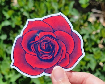 May include: A red and blue rose sticker with a white border. The sticker features a detailed illustration of a rose with red petals and blue shading, held against a backdrop of green foliage.