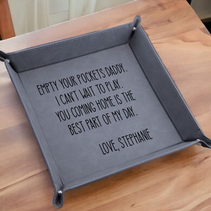 May include: A gray leather valet tray with the text "EMPTY YOUR POCKETS DADDY. I CAN'T WAIT TO PLAY. YOU COMING HOME IS THE BEST PART OF MY DAY. LOVE, STEPHANIE"
