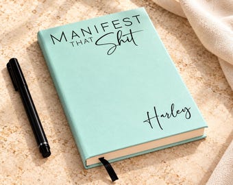 May include: A light mint green hardcover journal with a black ribbon bookmark. The cover features the black text "MANIFEST THAT SHIT" at the top and the name "Harley" in a script font at the bottom. A black pen lies next to the journal.