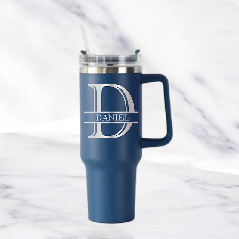 May include: A navy blue insulated tumbler with a handle and a clear lid. The tumbler features a large silver letter "D" with the name "DANIEL" in a horizontal bar across the middle. A clear straw is inserted into the lid.