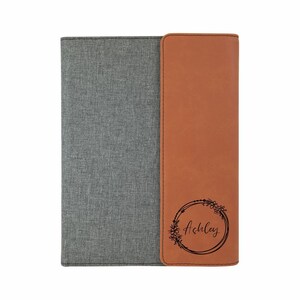 May include: A gray and brown leatherette notebook with a floral wreath design and the name "Ashley" engraved on the front.